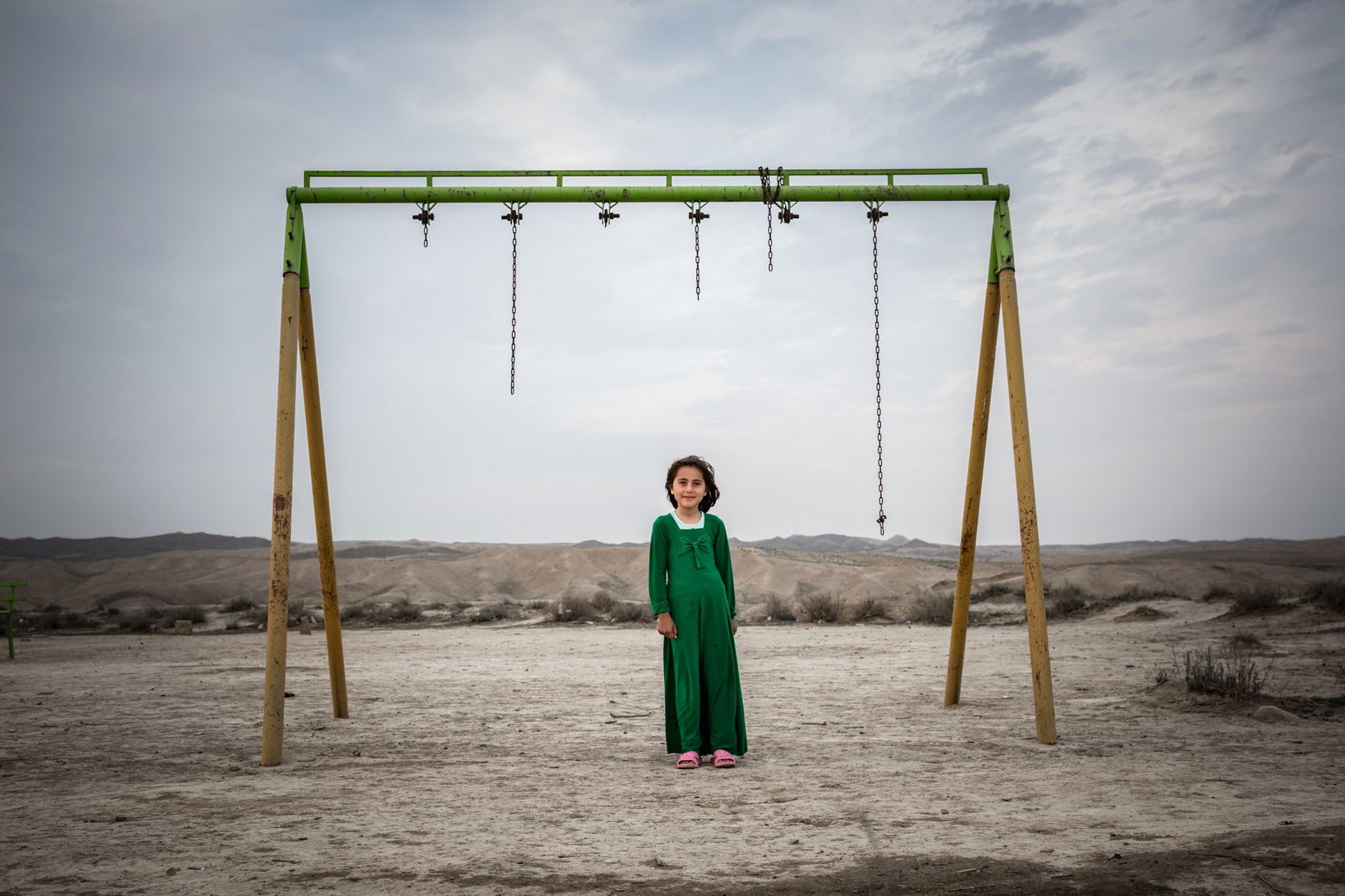 Portrait of a young girl stood in front of broken play swings with rugged desert landscapes in the background in Iran