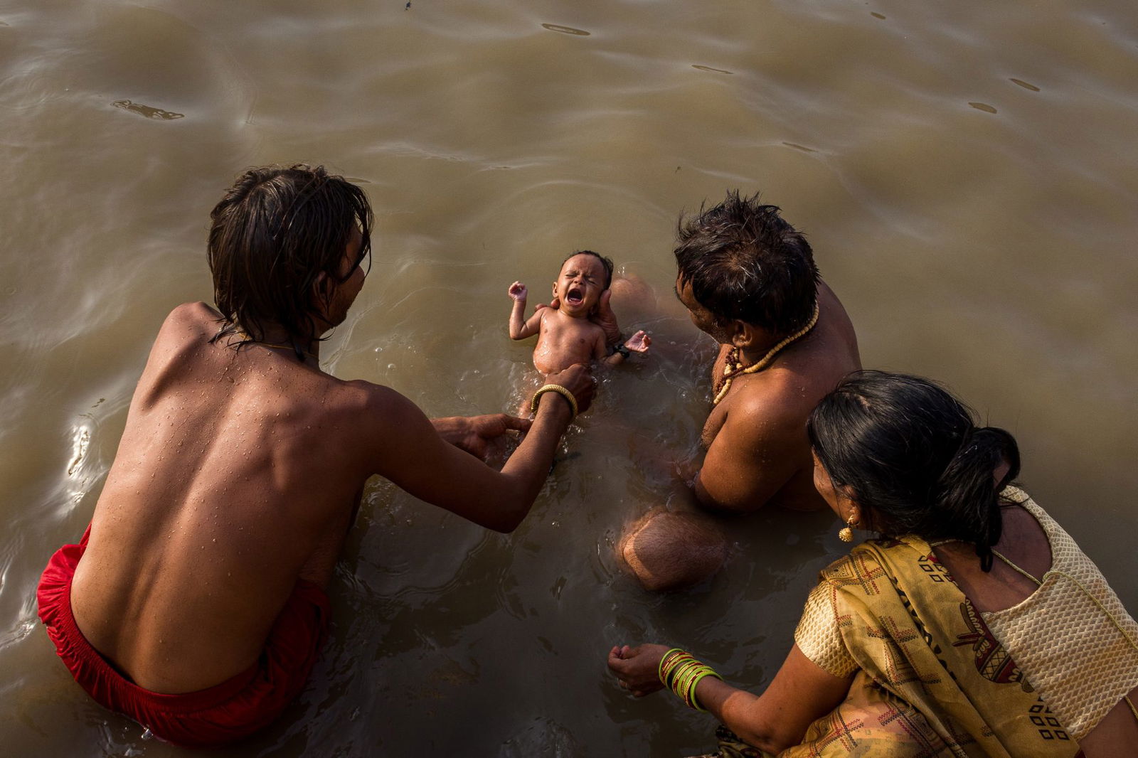 color photo of a family dipping their months old child in the Holy river Ganges -Varanasi, India by Mohit Khetrapal