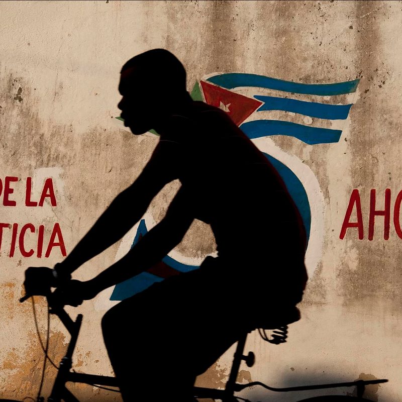 Color photo by Alex Almeida, boy cycling in front of cuban flag mural Cuba.