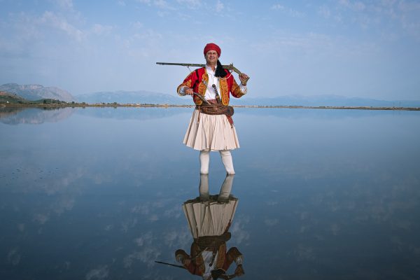 Color photo by Michael Pappas from Mitos - the thread of Greece, portrait - man in traditional costume with rifle, lake, mountains, sky