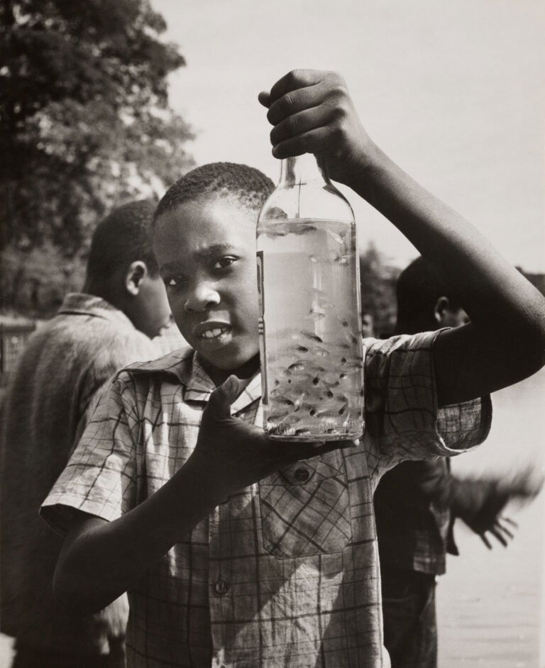 Street photography by Ralph Ellison. Black & white portrait of a young boy holding a bottle full of fish