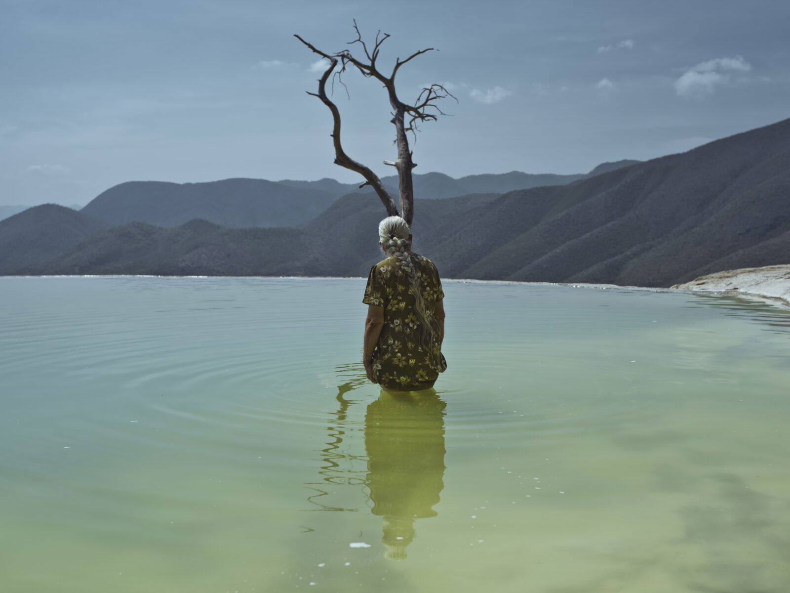 Photo of an elderly woman up to her waist in a lake surrounded by mountains by Cristina de Middel. From the book, Journey to the Center