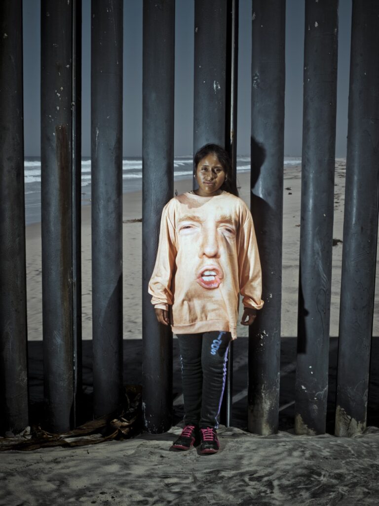 Portrait of a woman stood next to the US/Mexico border fence wearing a t shirt featuring Donald Trump's face by Cristina de Middel