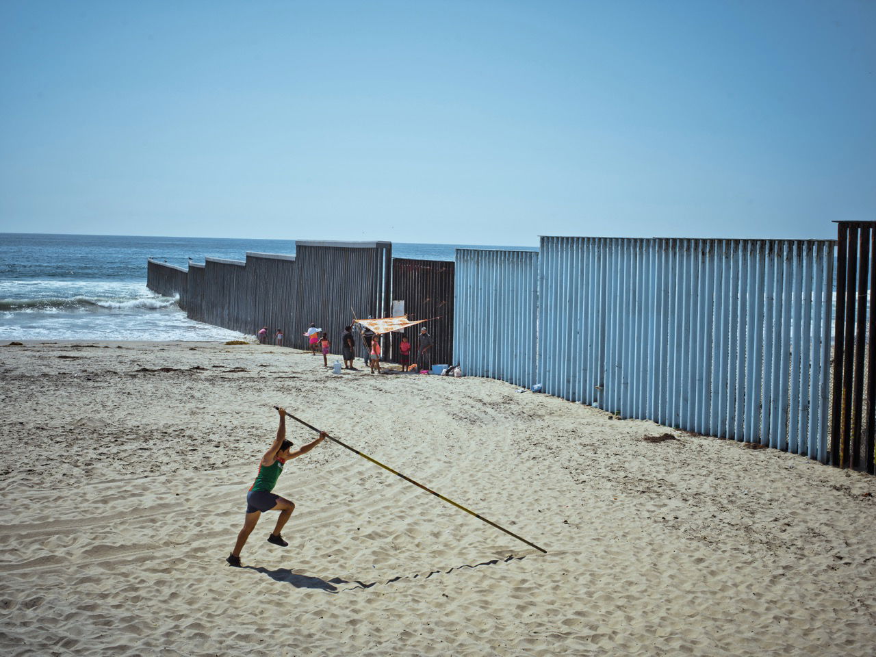 Photo of a man on a beach pole vaulting next to the US/Mexico border fence