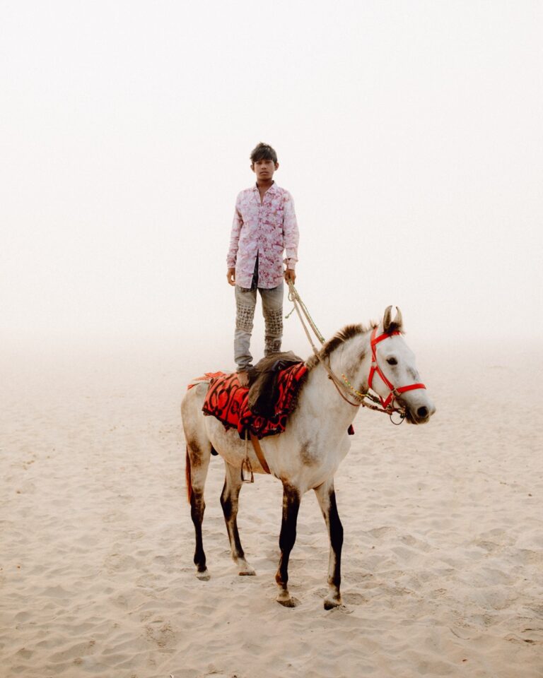 Photo of a boy stood on a horse in a sandy area by Pie Aerts