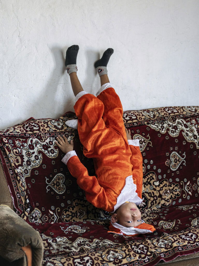 Documentary photography by Andrew McConnell. Portrait of a boy stood on his head on a sofa in Kenjebai-Samai, Kazakhstan