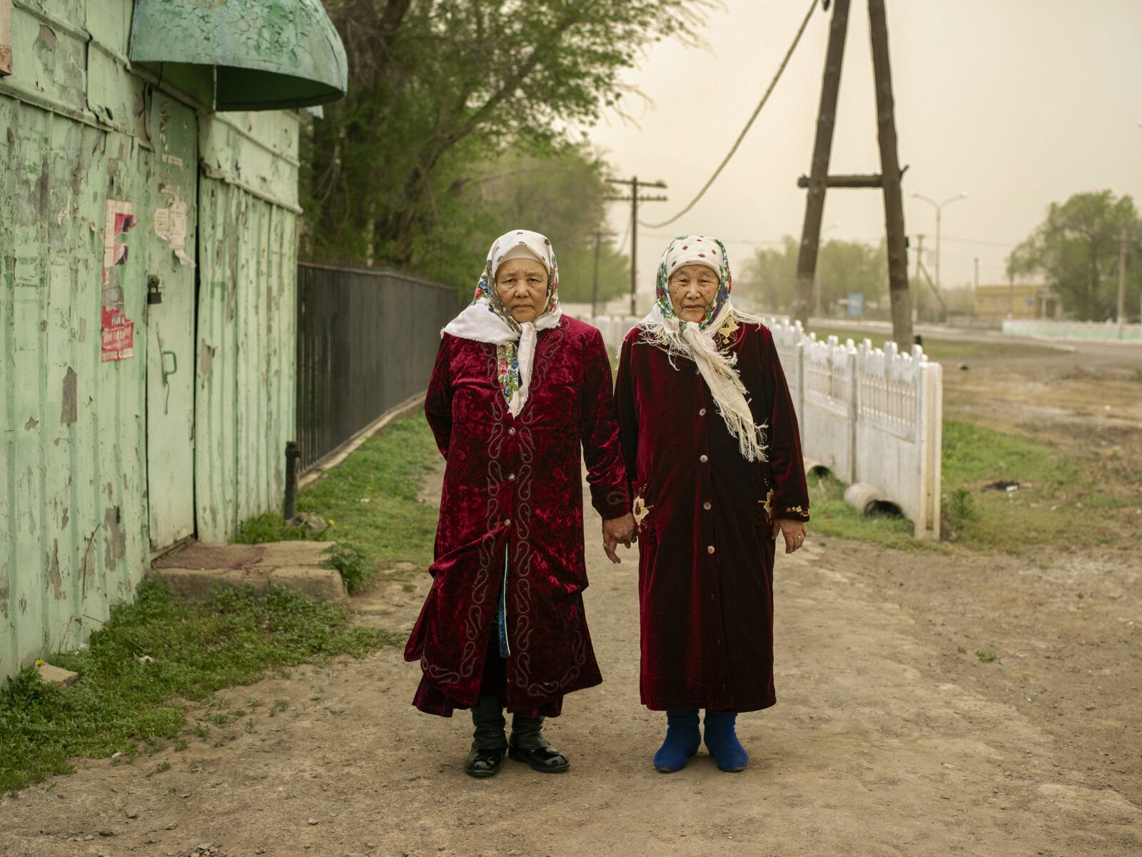 Documentary photography by Andrew McConnell. Two elderly women dressed in traditional clothing in front of a green building and some trees and dirt roads in Karaganda region, Kazakhstan, 2018