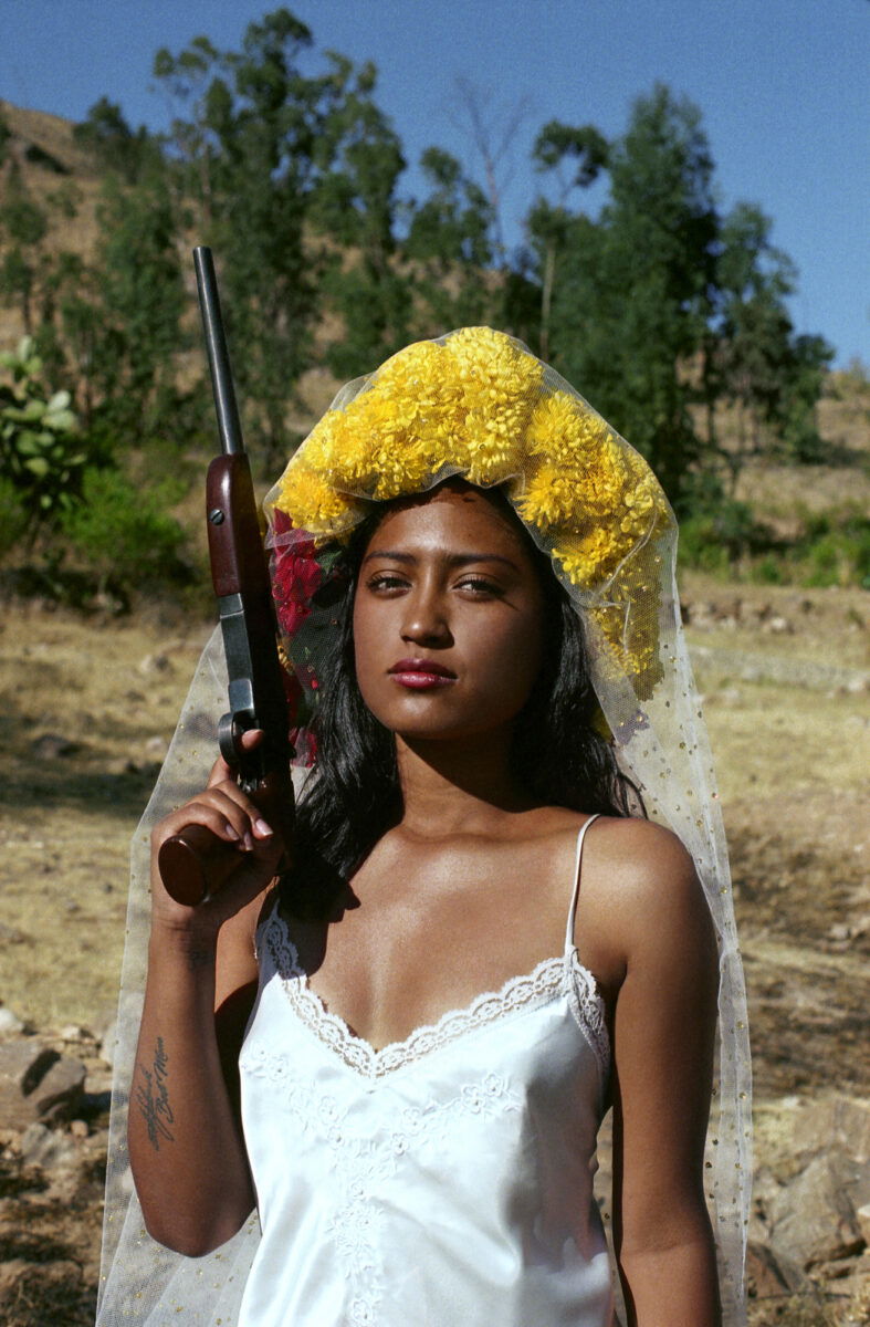 Portrait of a woman holding a rifle with yellow flowers on her head by Marisol Mendez